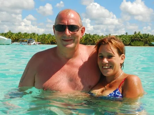 Manuela en Rainer, glimlachend in helder, turquoise water, met tropisch landschap op de achtergrond.