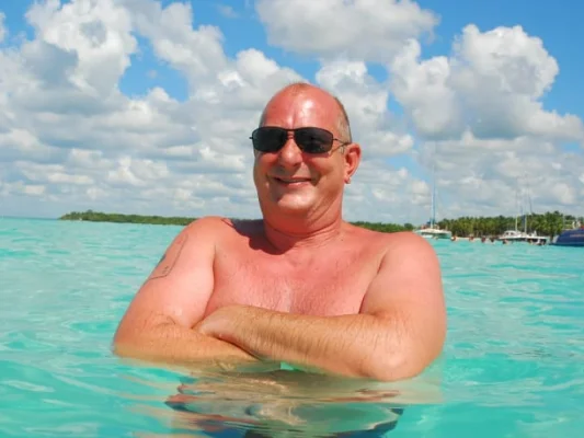 Rainer, glimlachend in turquoise water, met zonnebril en tropisch landschap op de achtergrond.