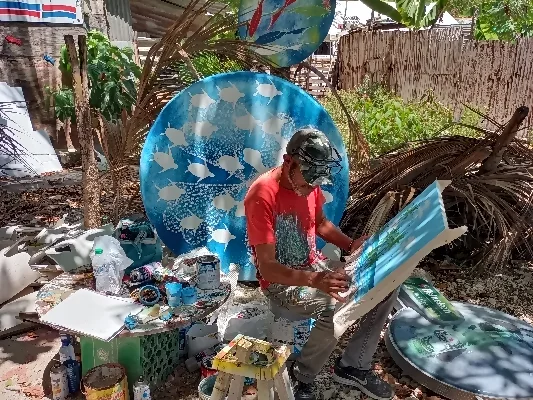 Kunstenaar in een openluchtatelier die aan een schilderij met maritieme motieven werkt, omringd door verschillende verfdozen en andere kunstwerken.