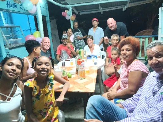 Groep mensen van verschillende leeftijden, waaronder kinderen, volwassenen en ouderen, zitten en staan rond een tafel bij een feestelijk buiten evenement met ballonnen en drankjes